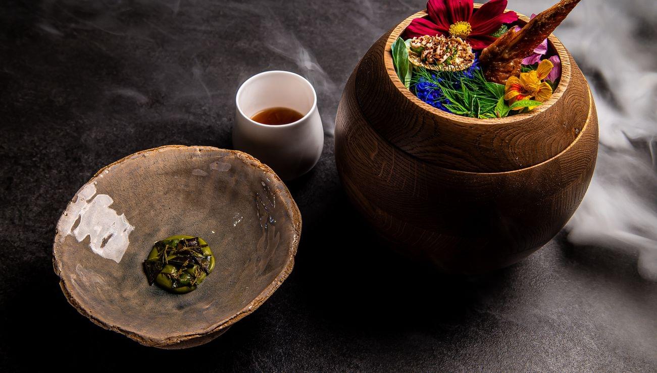 Gourmetgericht mit essbaren Blumen und Hähnchenflügel in Holzschüssel neben Keramiktellern auf dunkler Oberfläche