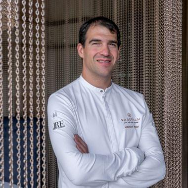Male chef in white uniform with Riederalm logo standing in front of decorative curtain