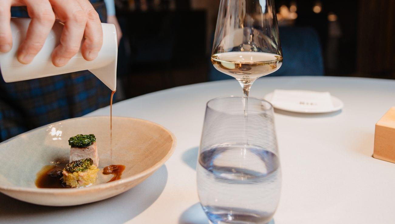 Gourmetgericht wird am Tisch mit Sauce verfeinert, dazu Wein- und Wasserglas auf elegantem Tisch