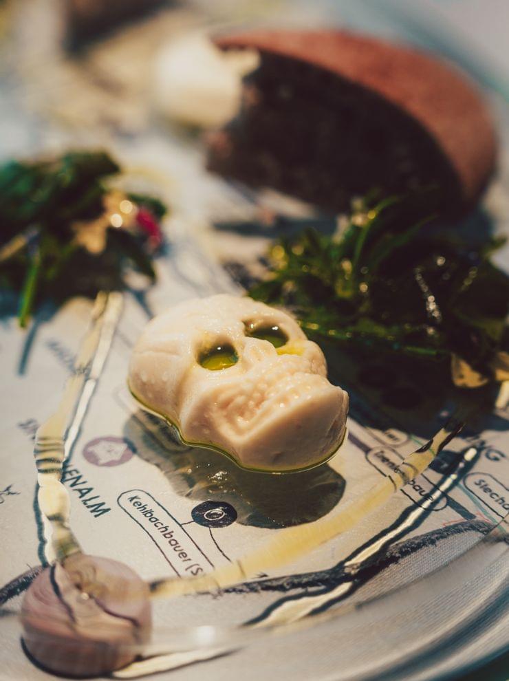 Gourmetgericht mit schädelförmiger Butter, dunklem Brot, Blattgemüse und Knoblauch auf dekorativem Teller