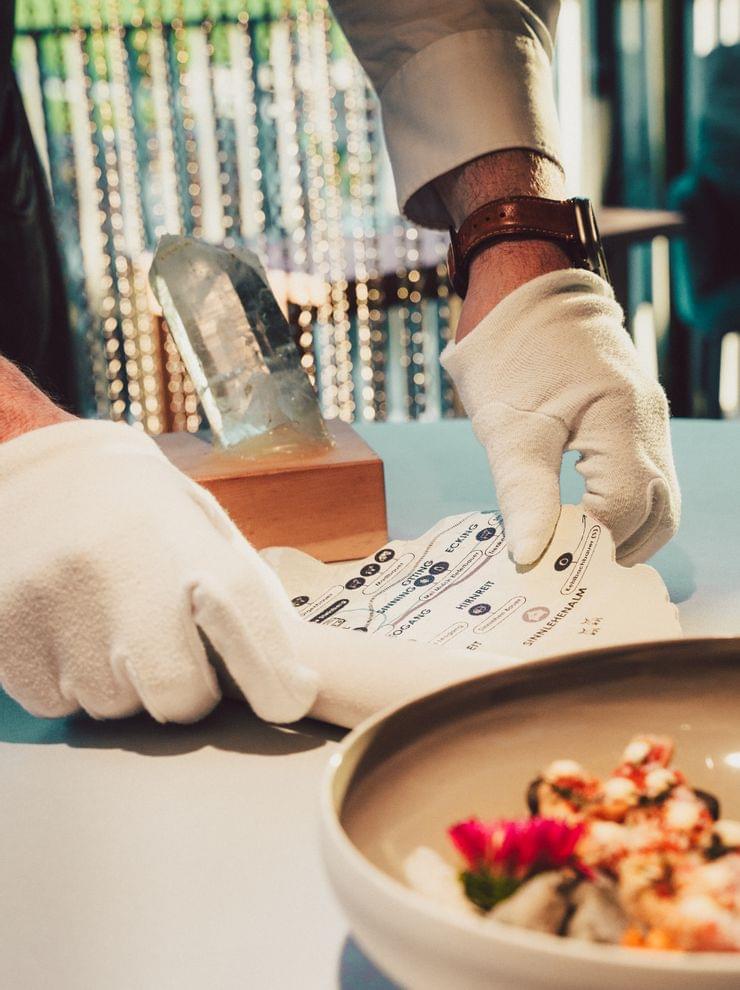 Behandschuhte Hände halten eine Speisekarte neben einem Gourmetgericht und Kristalldekor im modernen Restaurant