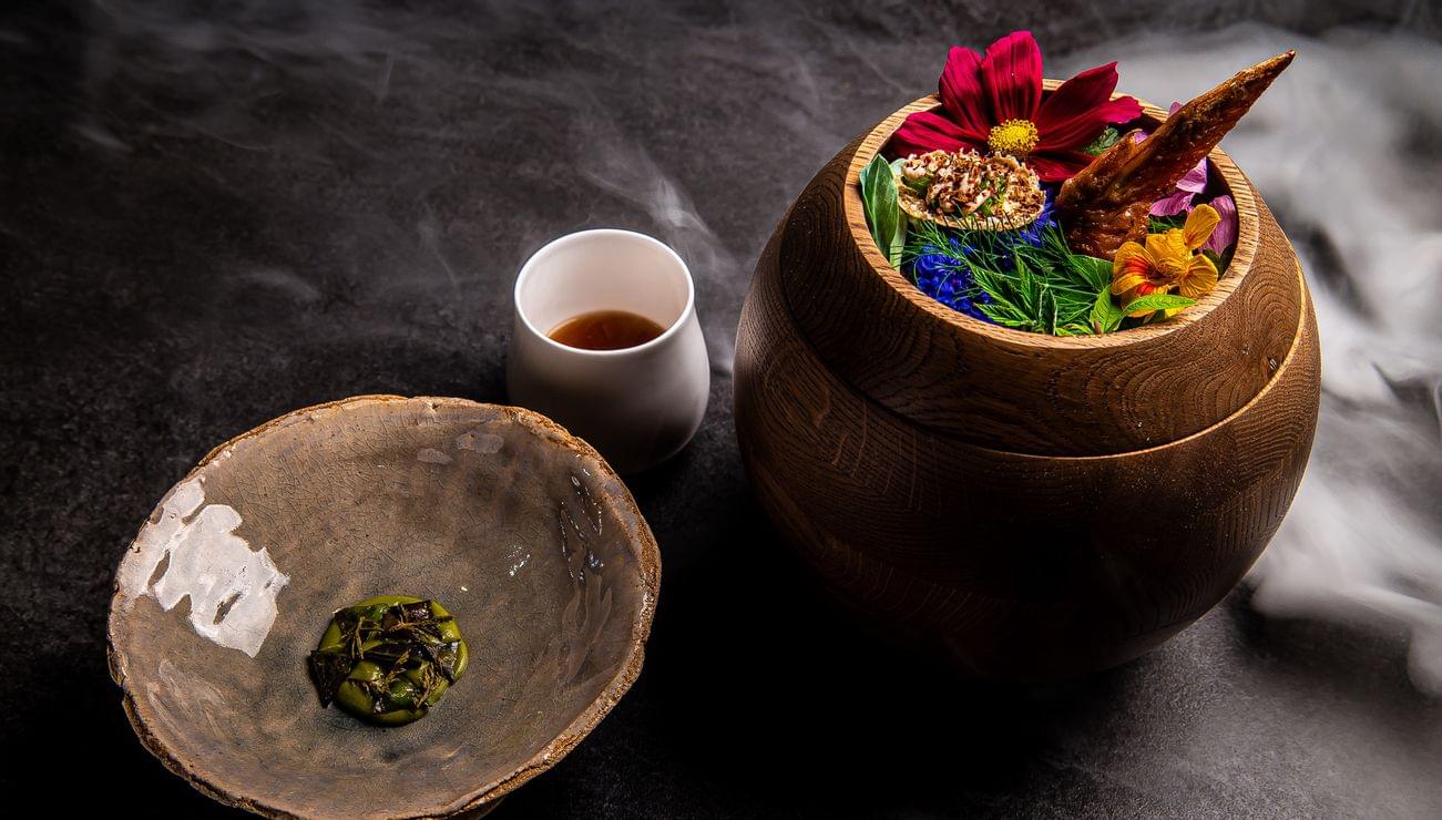 Gourmetgericht mit essbaren Blumen und Hähnchenflügel in Holzschüssel neben Keramiktellern auf dunkler Oberfläche